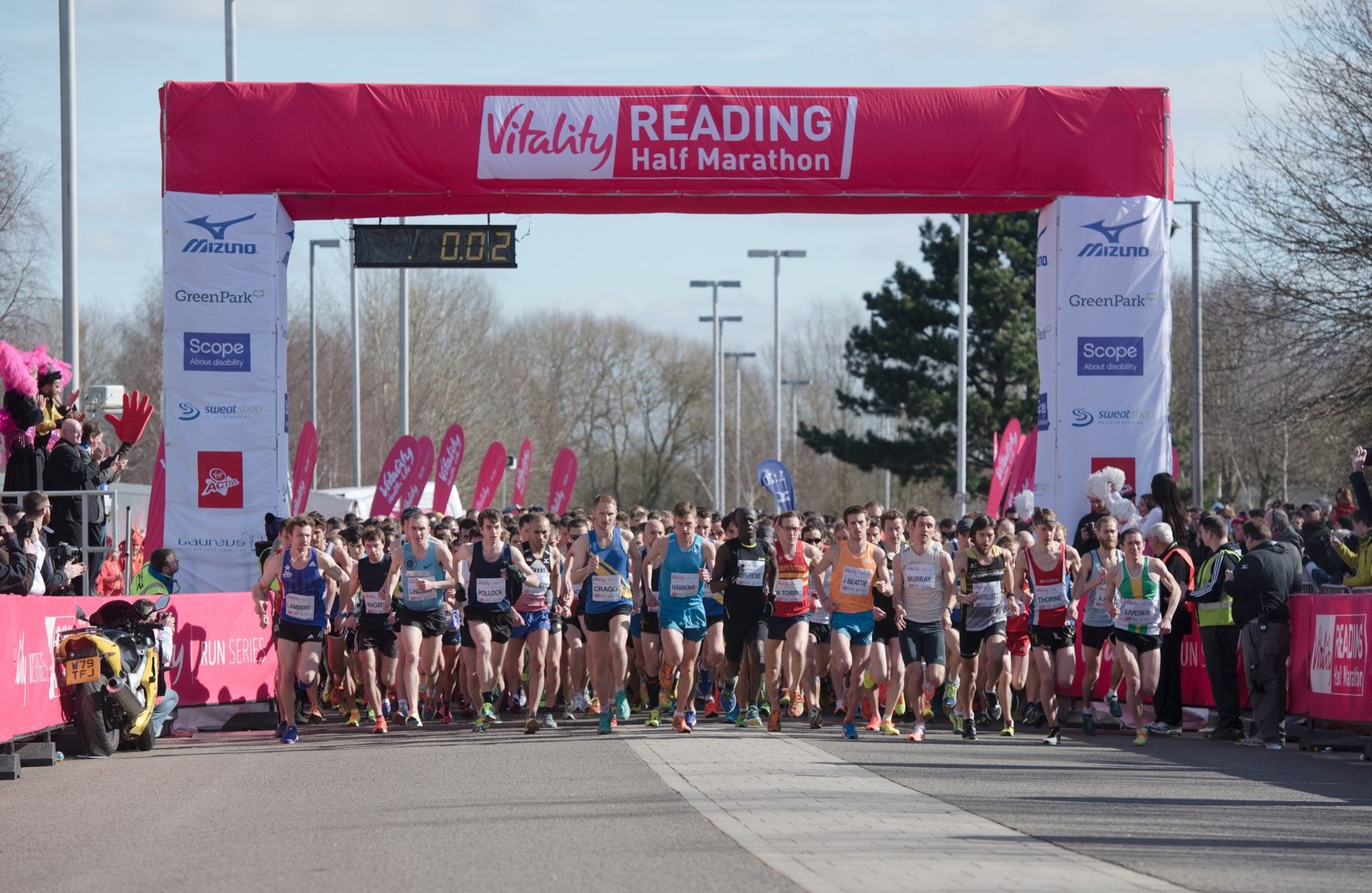 A large group of runners, wearing athletic gear, begin the Vitality Reading Half Marathon, passing under a pink and red starting arch. Spectators and officials stand on either side of the track, cheering. The timer above the arch shows "00:02". Trees and a clear sky are visible.