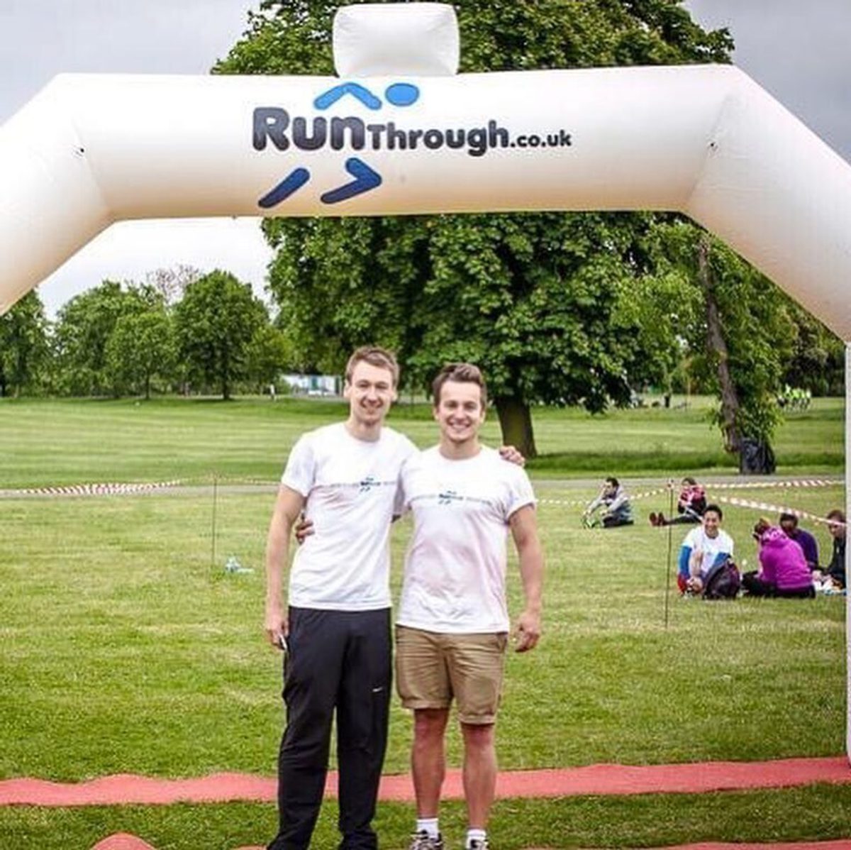 Two men dressed in athletic wear stand under a white inflatable arch with "Runthrough.co.uk" printed on it. They are smiling, standing on a grass field with trees in the background, and other participants resting on the ground. The event appears to be a race or fun run.