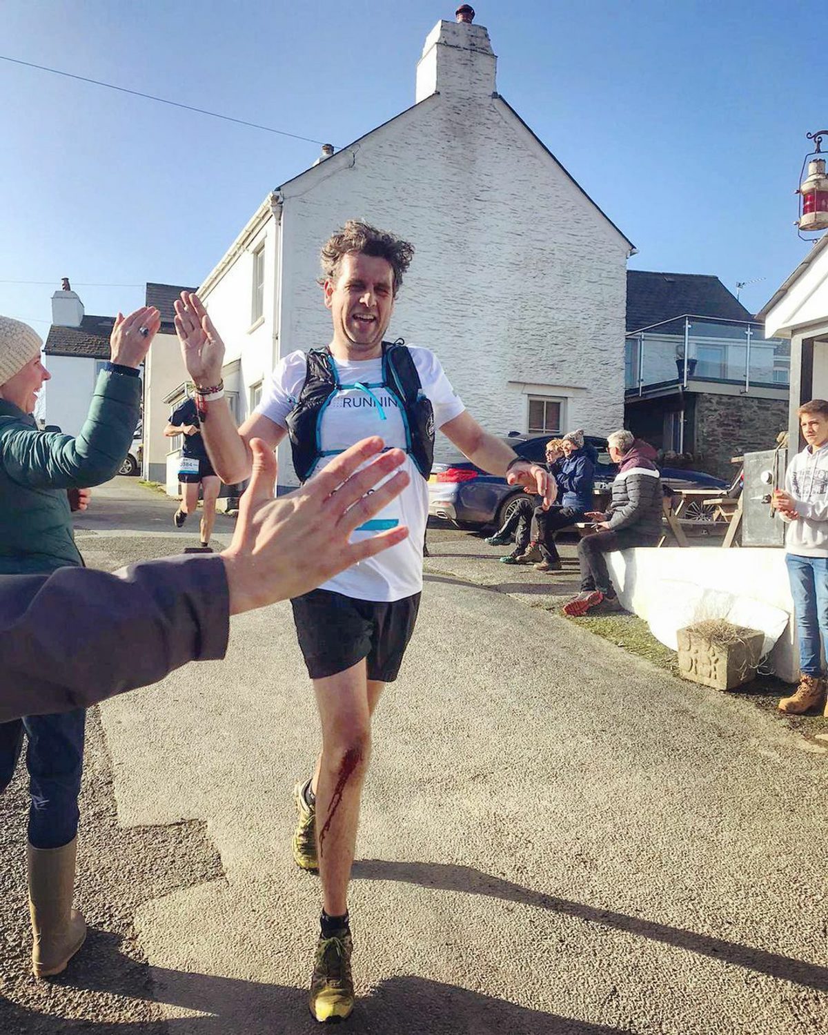 A runner wearing a white shirt and black shorts is high-fiving spectators as he finishes a race. He appears elated. The scene takes place on a sunny day in a village. People are cheering and sitting on benches near white houses in the background.