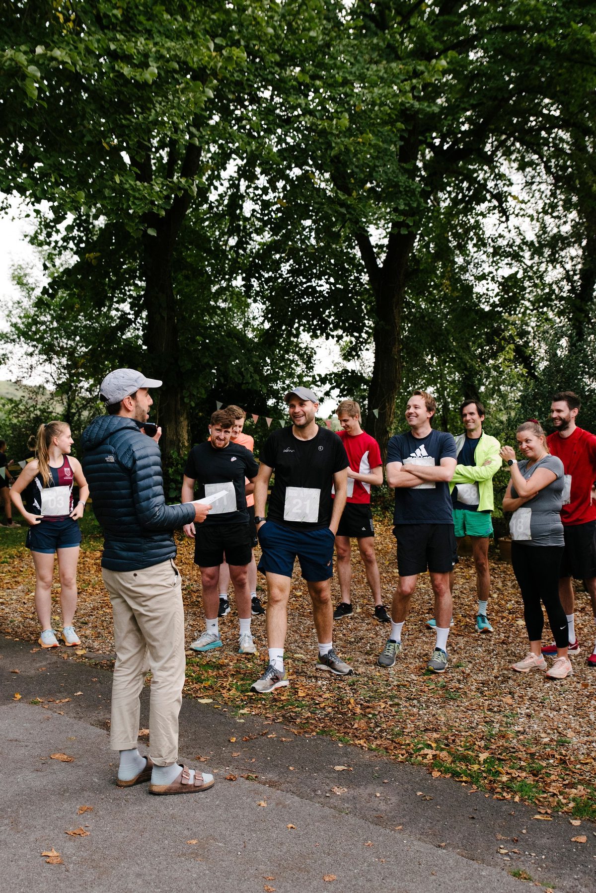A group of runners stands in a park, attentively listening to a man in a cap and jacket who appears to be giving instructions. The runners are dressed in athletic wear, some with race numbers pinned to their shirts. Trees and foliage surround them.