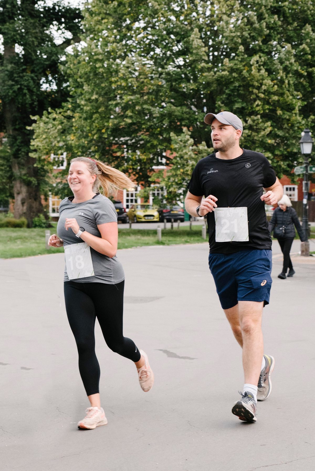 A man and a woman, both wearing numbered race bibs (#21 and #18), are running together on a paved path in a park. The woman is on the left wearing a gray shirt and black leggings, while the man is on the right wearing a black shirt and navy shorts. Trees and buildings are visible in the background.