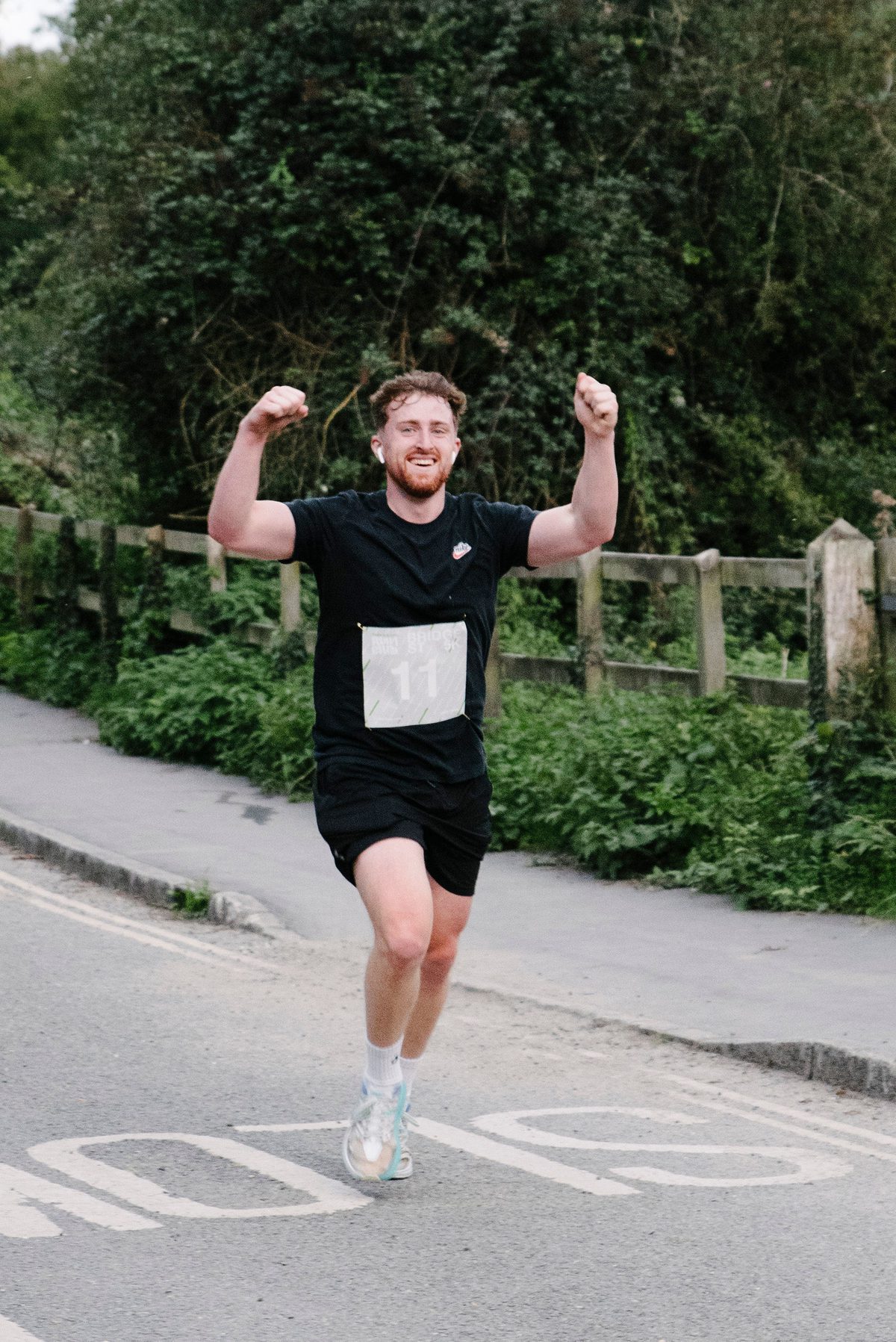 A man in a black T-shirt and shorts, wearing a race bib with the number 11, is running on a road with arms raised in a victory pose. He is smiling, and there is greenery and a wooden fence in the background.