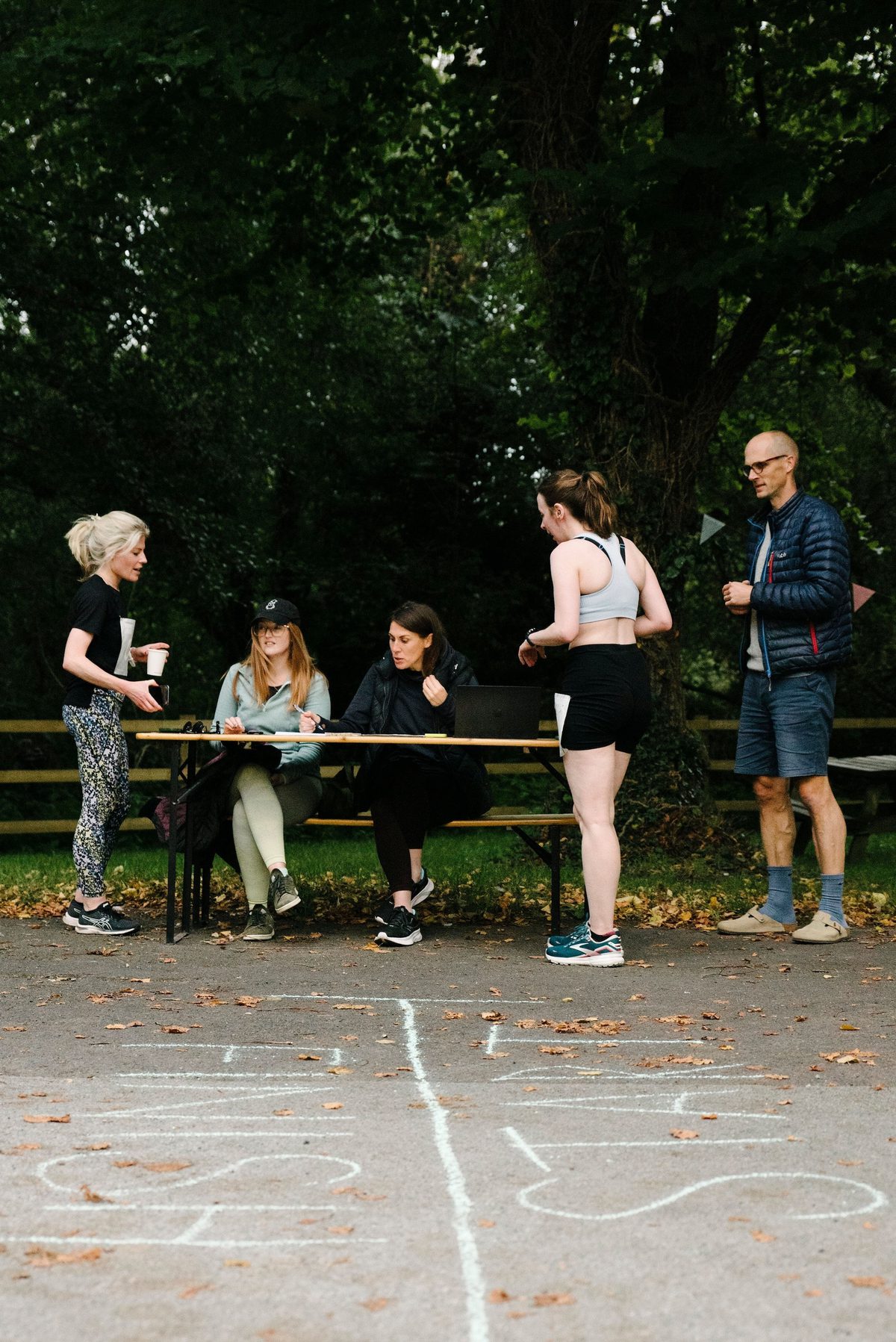 Four people are gathered around a small table outdoors, with two women seated and two women standing. An older man stands nearby, looking on. The ground in front has white chalk markings. The scene is relaxed, set in a leafy park area.
