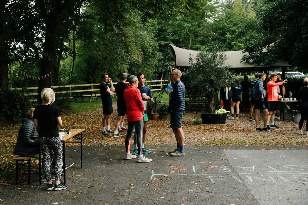 A group of people stands in a park area, conversing and preparing for an outdoor activity or event. Some are dressed in athletic wear, while others are talking near a registration table under a canopy. The ground is covered with fallen leaves, and the area is surrounded by trees.