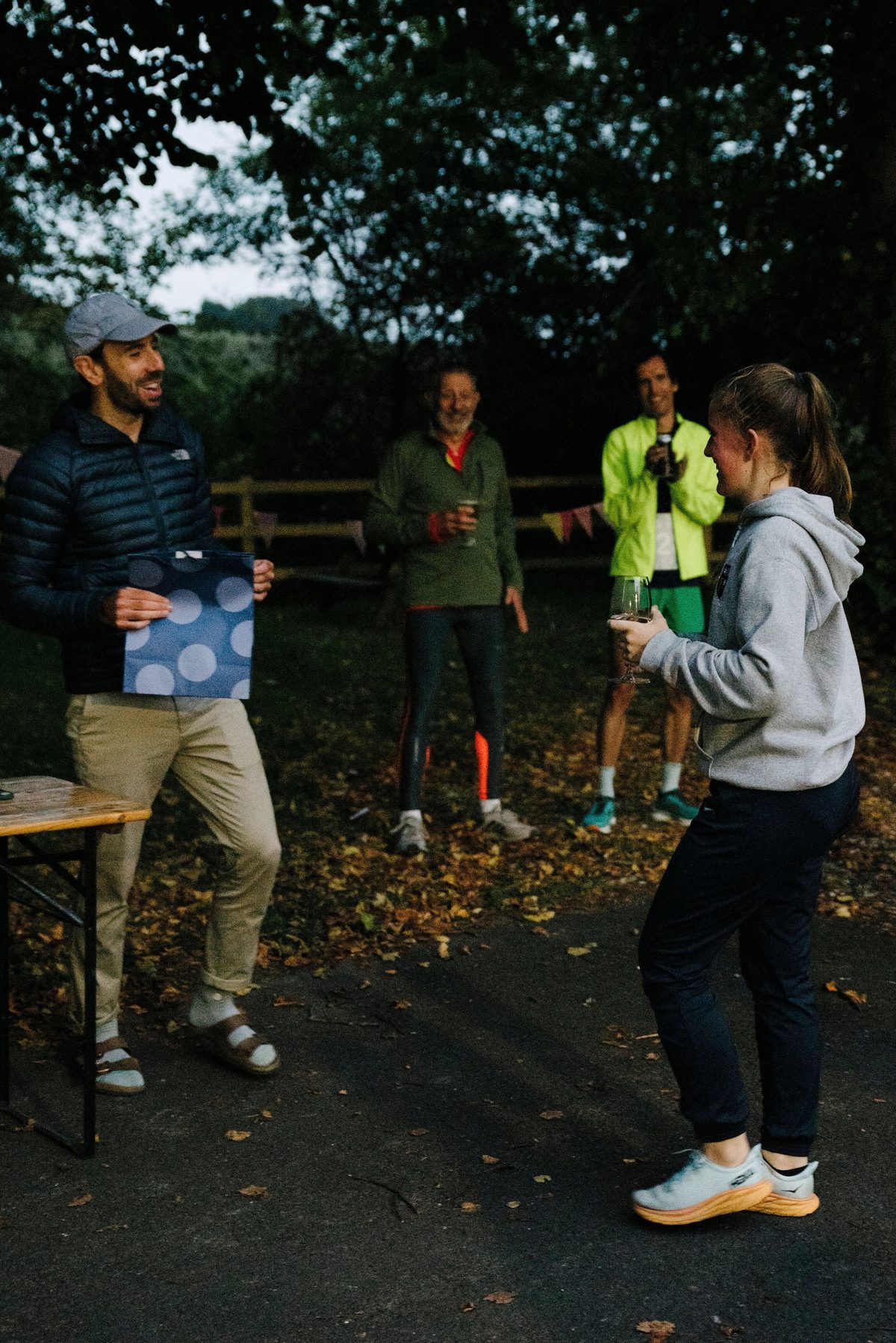A person in a hat and jacket smiles while holding a polka-dotted piece of paper, standing near a person in a hoodie holding a cup. Two others in bright athletic clothing stand in the background, smiling. They are outside on a path surrounded by trees and fall leaves.
