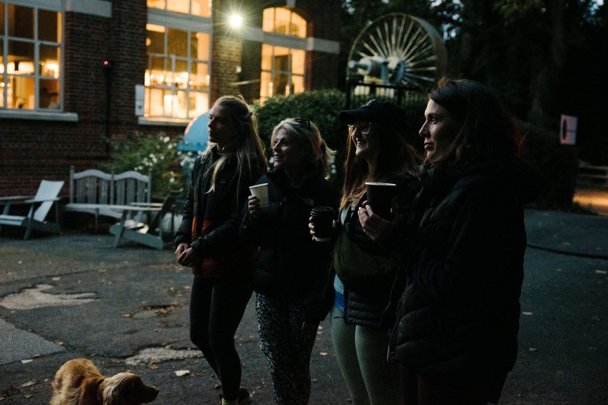 Four women stand outdoors in the early evening, facing left. One holds a coffee cup, and a golden retriever sits at their feet. They are dressed warmly in jackets and hoodies. Behind them is a lit brick building with large windows and outdoor seating.