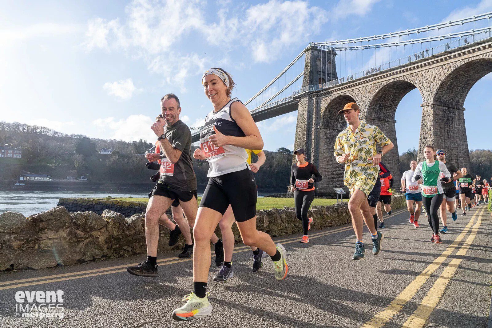 A group of runners participates in a race on a sunny day near a scenic bridge. They run on a paved road, and the backdrop features a river, bridge, and green hills. They are dressed in athletic attire, and some wear race bibs. The mood is energetic and focused.