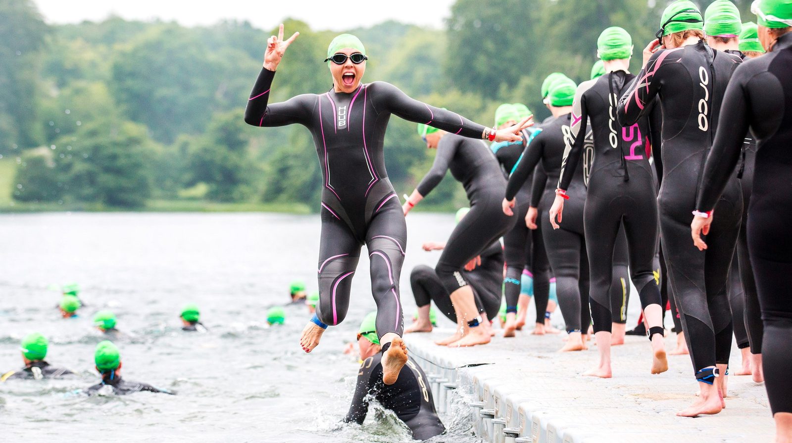 A group of swimmers in wetsuits and green swim caps are on a dock by a lake, preparing to enter the water. A joyful swimmer in the foreground, with arms raised in excitement, jumps off the dock into the lake. The background shows a green, wooded area.