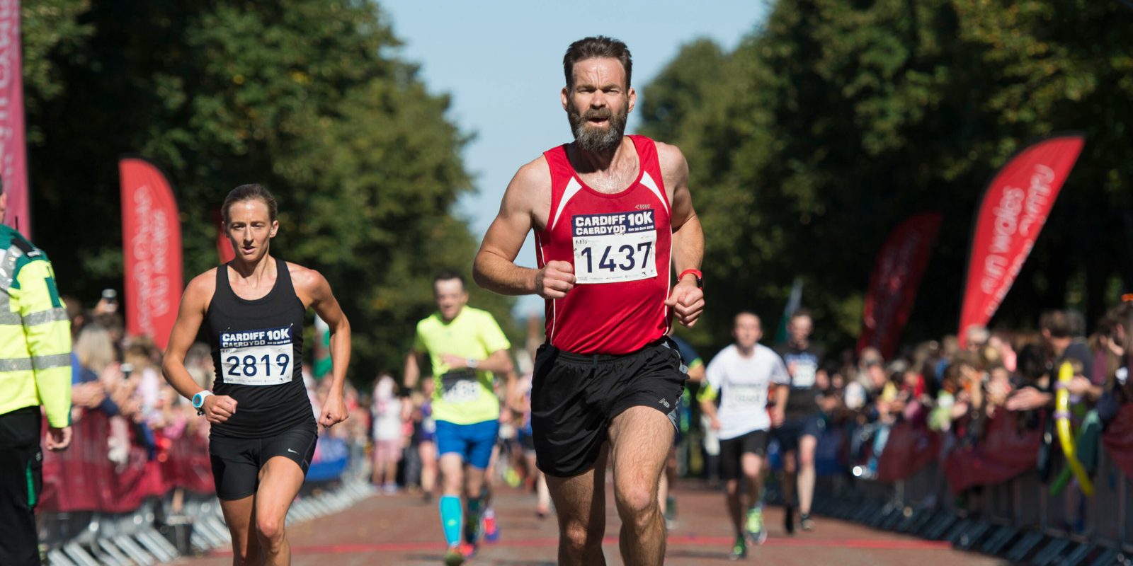 Runners participate in the Cardiff 10K race. A man with a beard and red shirt, bib number 1437, leads while others follow closely. Trees and cheering spectators line the path. The event appears to take place on a sunny day.