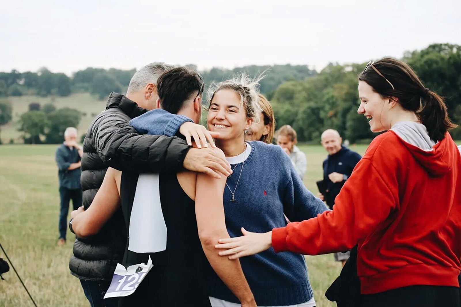 A group of people are gathered outdoors, smiling and embracing each other. They appear to be celebrating. One person is wearing a sports bib with the number 12. The background shows a grassy field and trees. The mood is joyful and congratulatory.
