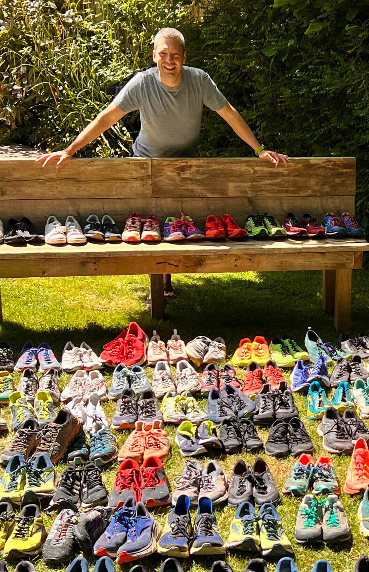 A person stands behind a wooden bench in a grassy yard, smiling with hands on the bench. In front of the bench, numerous pairs of colorful running shoes are neatly arranged in rows on the ground, organized by different styles and colors.