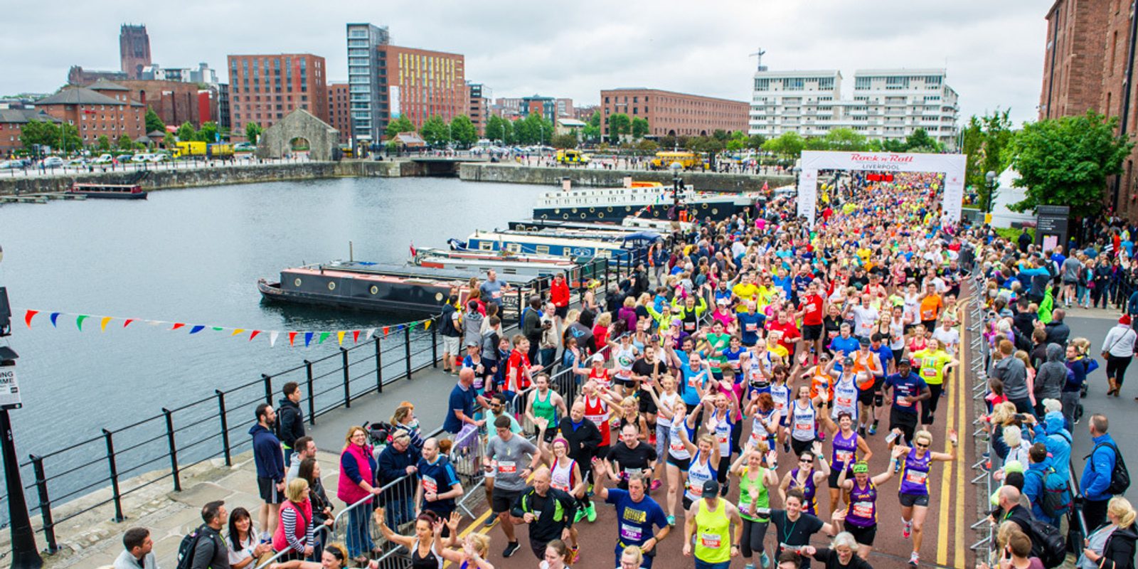 A large crowd of runners, wearing various colorful outfits, participates in a marathon along a waterfront with boats docked. Spectators line both sides of the path, and buildings are visible in the background. The sky is overcast.