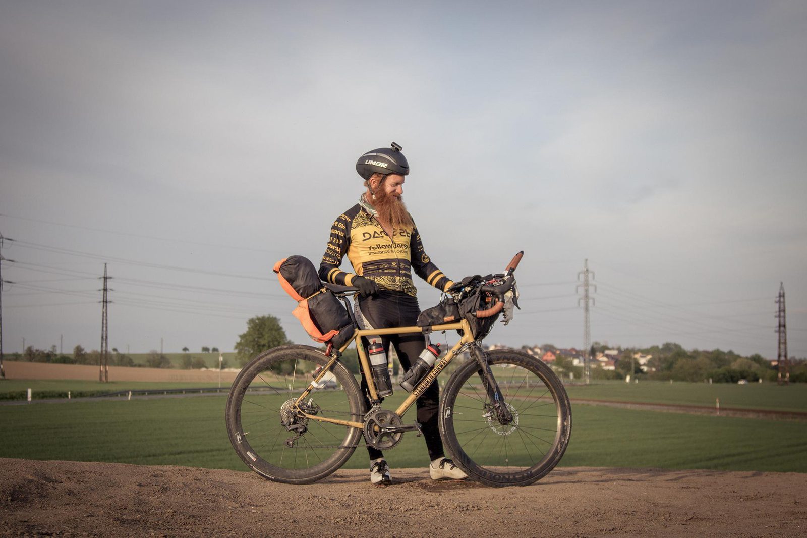 A cyclist with a long beard stands beside a loaded touring bike in a rural landscape. The bike has bags attached to the front and rear. The cyclist wears a helmet, sunglasses, and a yellow and black jersey. Farmland and power lines are visible in the background.