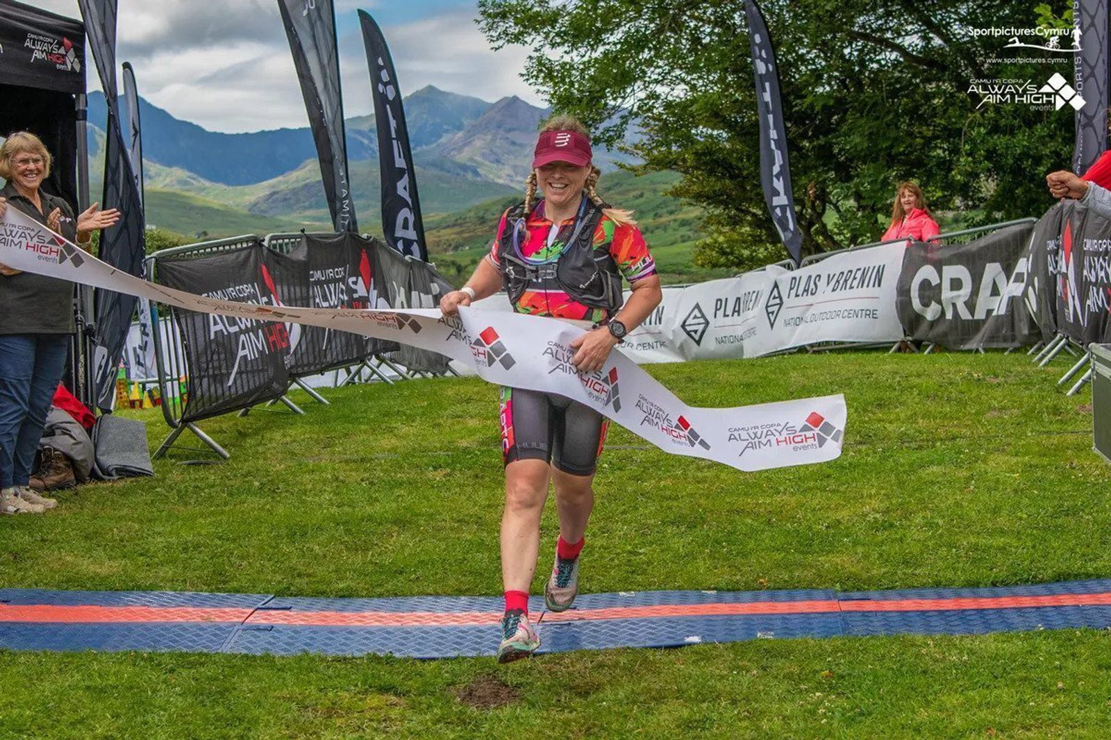 A person wearing a triathlon suit and cap is crossing a finish line ribbon with a smile. A cheering crowd is visible behind barricades. The backdrop features mountains, lush greenery, and event banners. The sky is partly cloudy.