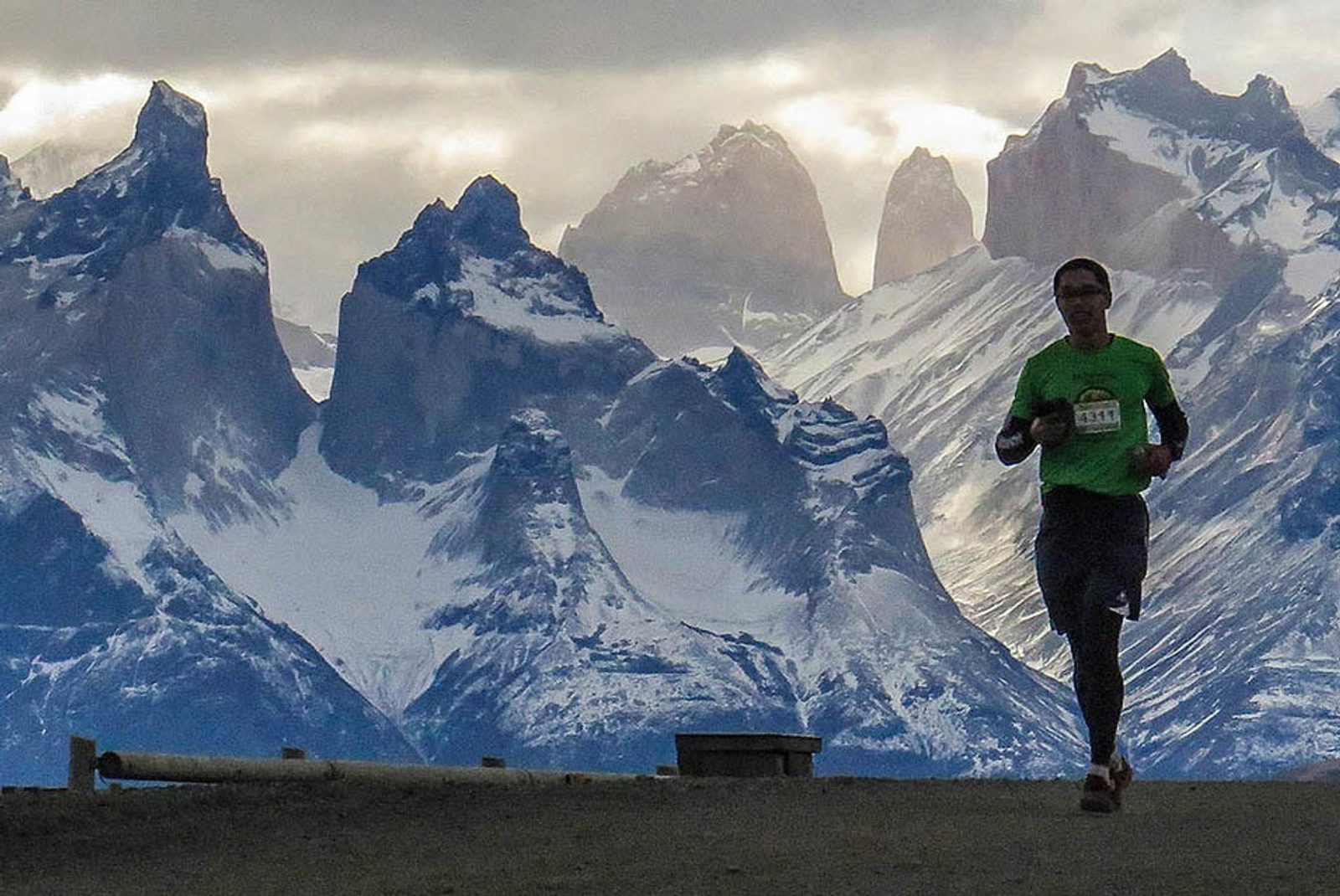 A runner in a green shirt and black pants jogs on a trail with a dramatic backdrop of snow-covered, jagged mountains under a cloudy sky. The runner is holding a bottle and appears to be participating in a race. The scenery is rugged and awe-inspiring.