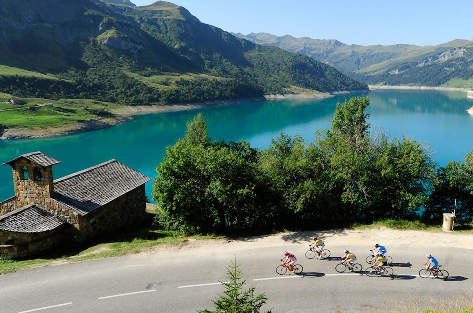 Cyclists ride along a scenic road with a turquoise lake and lush green mountains in the background. An old stone cabin with a slanted roof is situated near the edge of the lake, surrounded by trees and natural beauty. The sky is clear and sunny.
