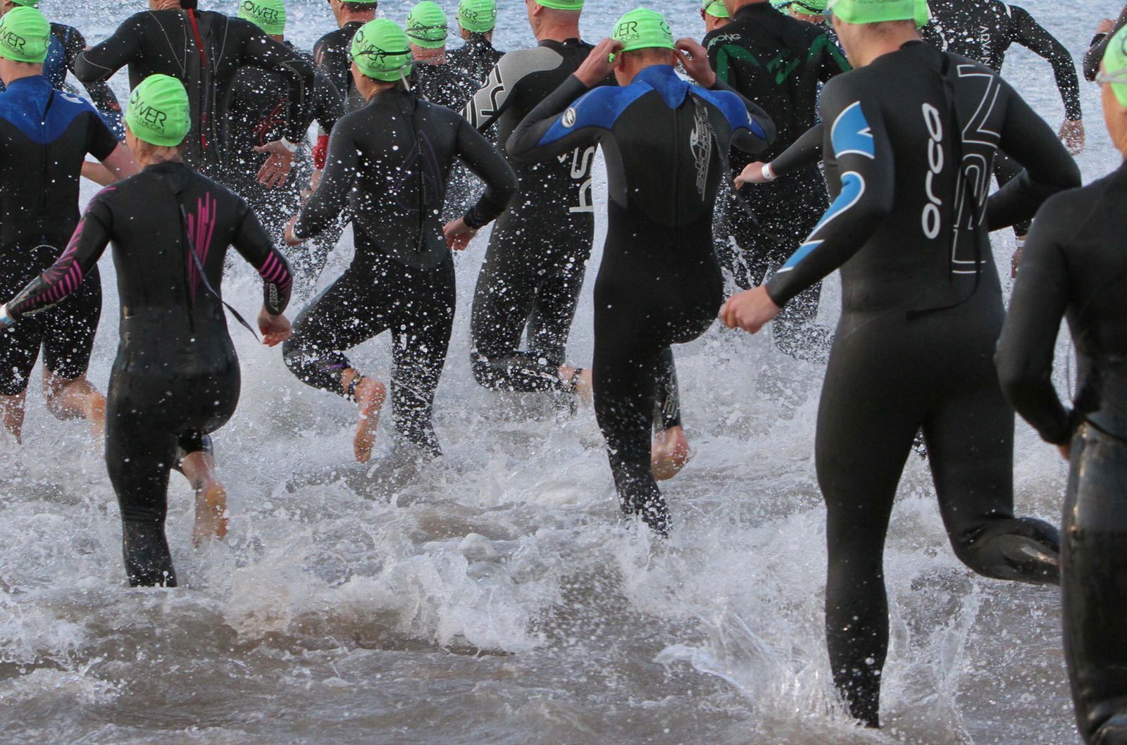 A group of athletes wearing black wetsuits and green swim caps run into the ocean, splashing water around as they begin a triathlon or open water race. The image captures the moment from behind, showing the energy and movement of the competitors.