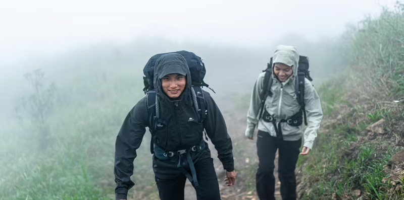 Two individuals, dressed in weatherproof jackets with hoods, hike up a foggy trail. They are both carrying backpacks and appear to be enjoying their adventure despite the misty conditions. The scene is lush with green vegetation on either side of the trail.