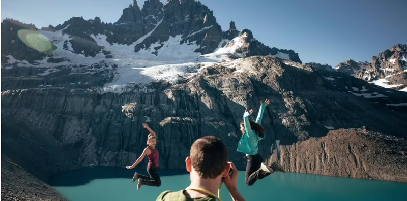 Three people jump in the air in front of a stunning mountain landscape with snow patches and a turquoise lake below. One person faces away, capturing the scene with a camera, while the other two face the camera, arms extended in celebration and joy.