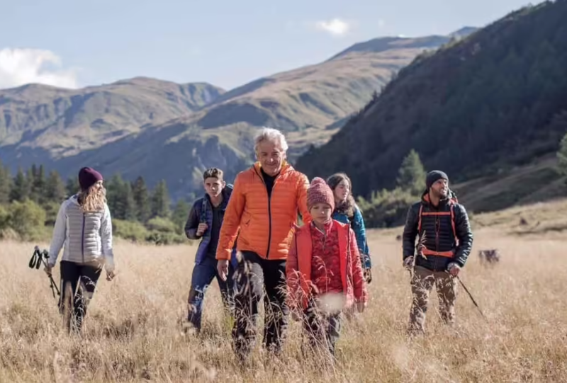 A group of six people, including an older adult, children, and several adults, are hiking through a grassy field with mountain scenery in the background. They are all dressed in warm jackets and hats, suggesting a cool climate. They appear to enjoy their outdoor adventure.