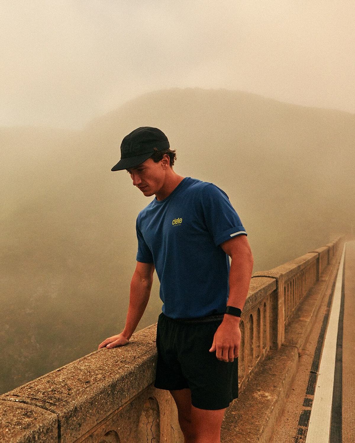 A person wearing a blue shirt, black shorts, and a black cap is standing on a bridge with a foggy landscape in the background. They are looking down and touching the bridge railing with one hand.