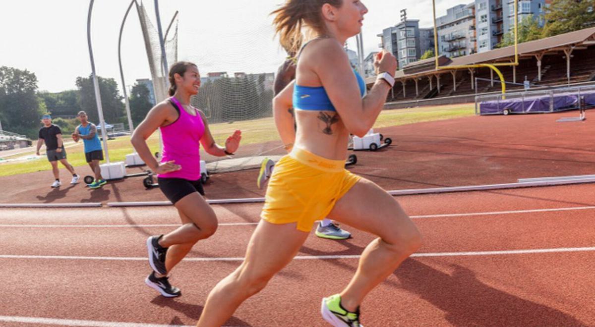 Three runners on an outdoor track, with a woman in yellow shorts and a blue sports top leading the group. Behind her, a woman in a pink tank top and black shorts, and another person in green are running. Buildings and sports equipment can be seen in the background.