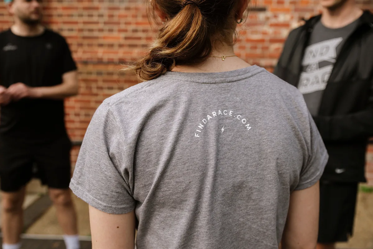 A woman with her back to the camera is wearing a grey T-shirt with "FINDARACE.COM" written on it. She has a ponytail and appears to be talking to two people in casual athletic wear in front of a brick wall.