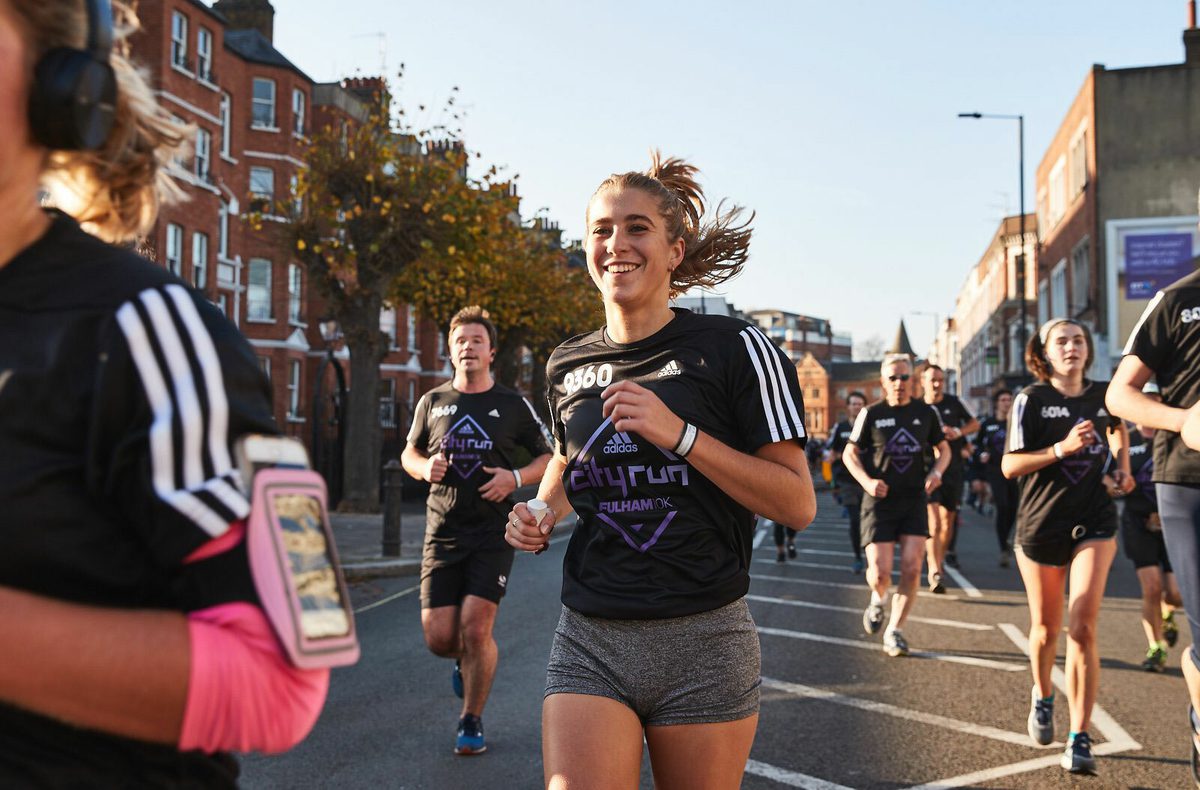 A group of runners in athletic wear participate in a road race on a city street. The front runner, a woman with a ponytail, looks happy as she runs. Other runners are spread out behind her. A row of buildings and autumn foliage are visible in the background.