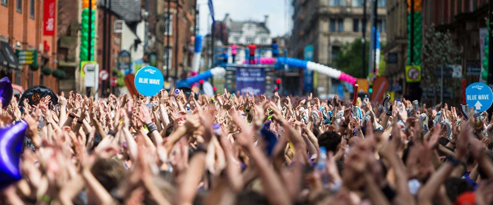 A large, energetic crowd of people with hands raised, filling a city street lined with buildings. Blue and pink balloons and arches can be seen in the background, along with colorful signage and decorations.
