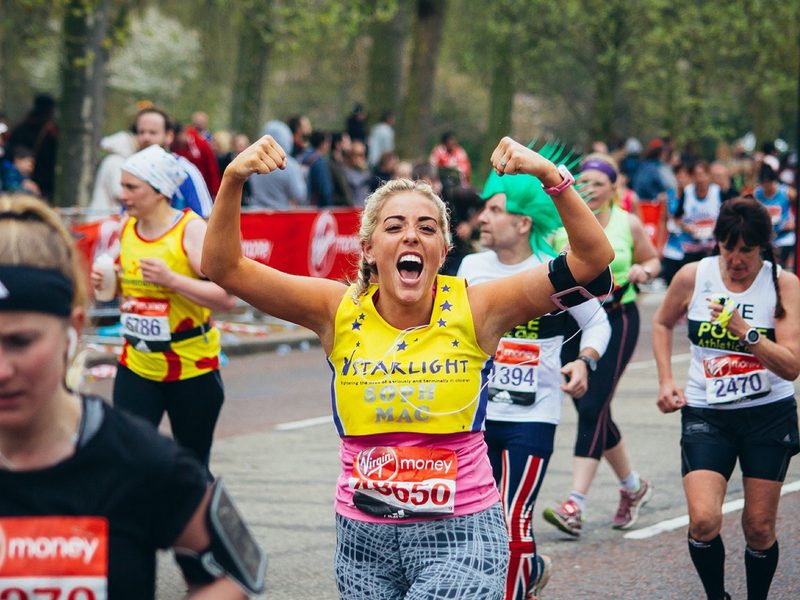 A woman with blonde hair, wearing a yellow "Starlight" tank top and patterned leggings, excitedly flexes her arms while running in a race. Other runners are visible in the background, participating in the event in a park setting.