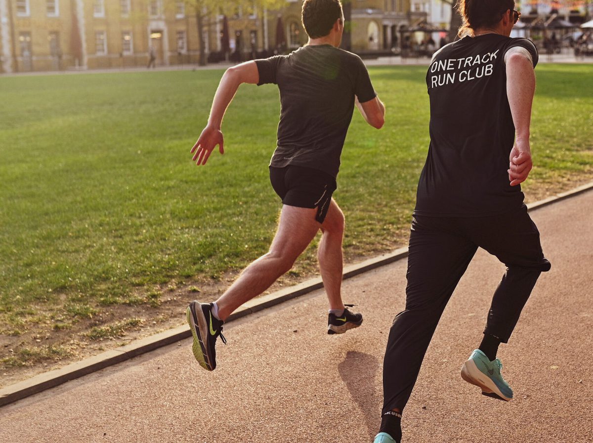 Two people in athletic gear are running on a paved path beside a grassy area in a park. One person wears a black T-shirt with "Onetrack Run Club" written on the back. The sun is setting, casting a warm light over the scene.