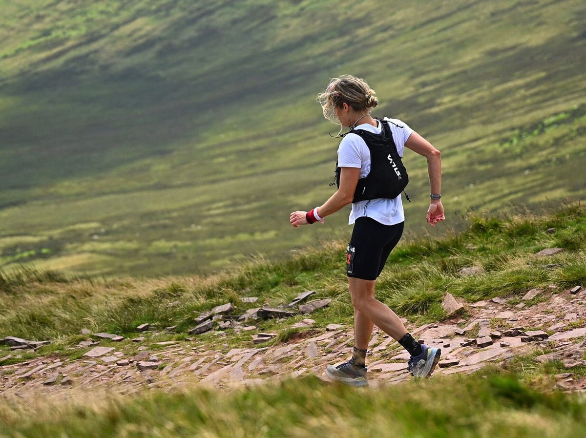 A person with light hair in a ponytail, wearing a white shirt, black shorts, and a running vest, walks on a rocky trail through grassy hills in a rural outdoor landscape.