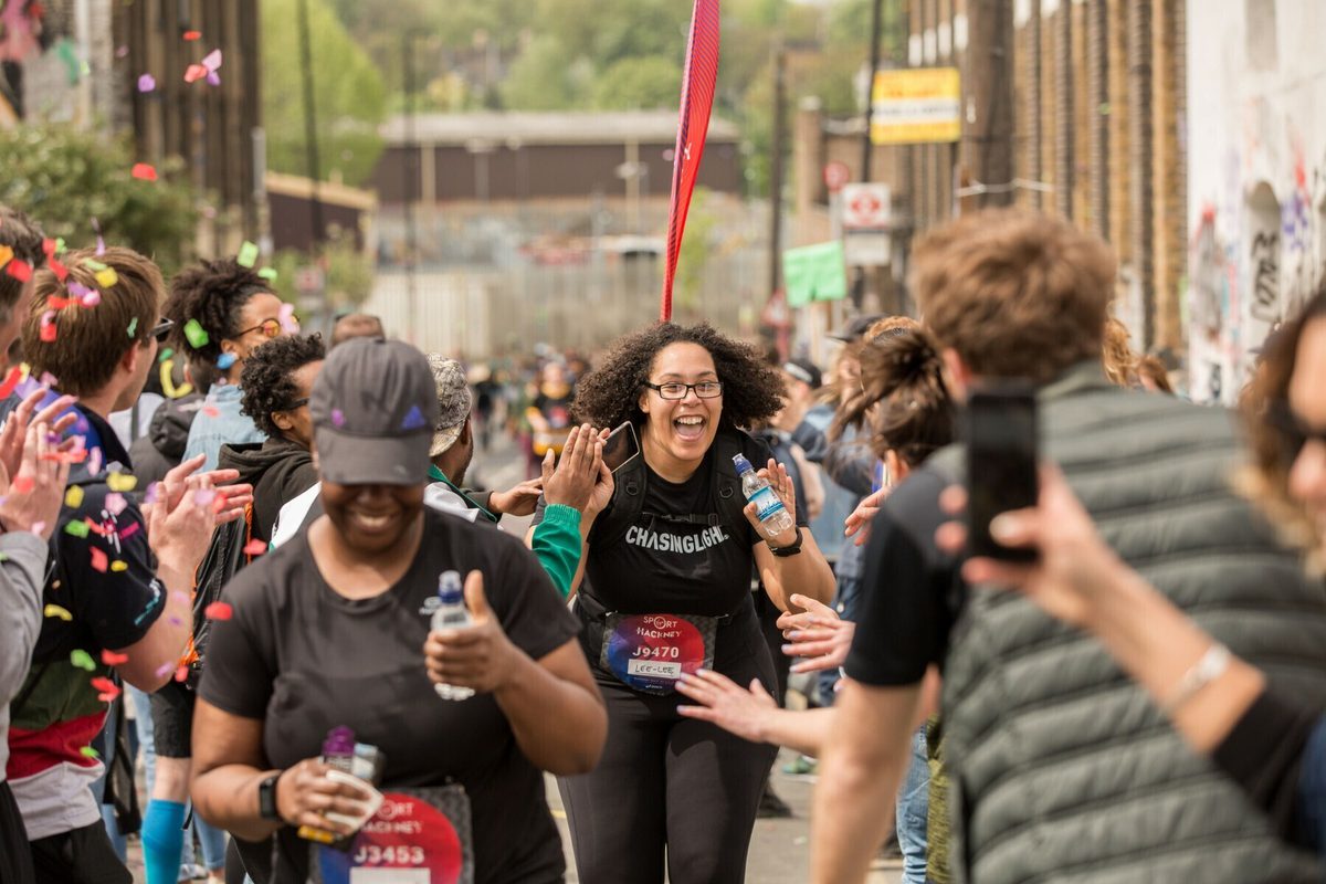 A woman joyfully runs through a crowded street during a race, with onlookers cheering her on and confetti flying in the air. She wears a black shirt that says "CHASING". Other runners and spectators surround her, creating an energetic atmosphere.
