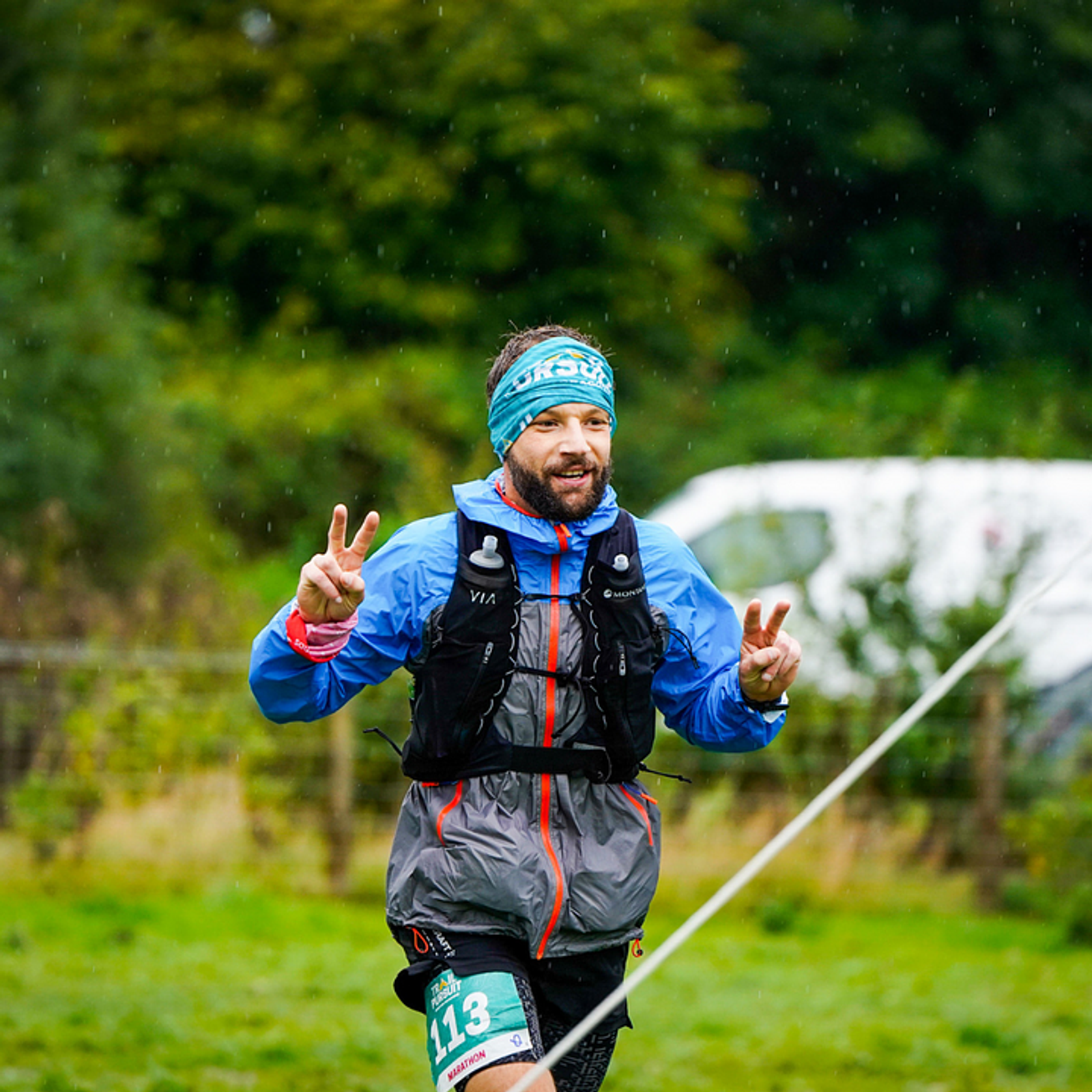 A runner in outdoor athletic gear, including a blue jacket and a headband, is making peace signs with both hands while running. He appears to be participating in a race, with a green race bib numbered "113" and a grassy background. It is raining slightly.