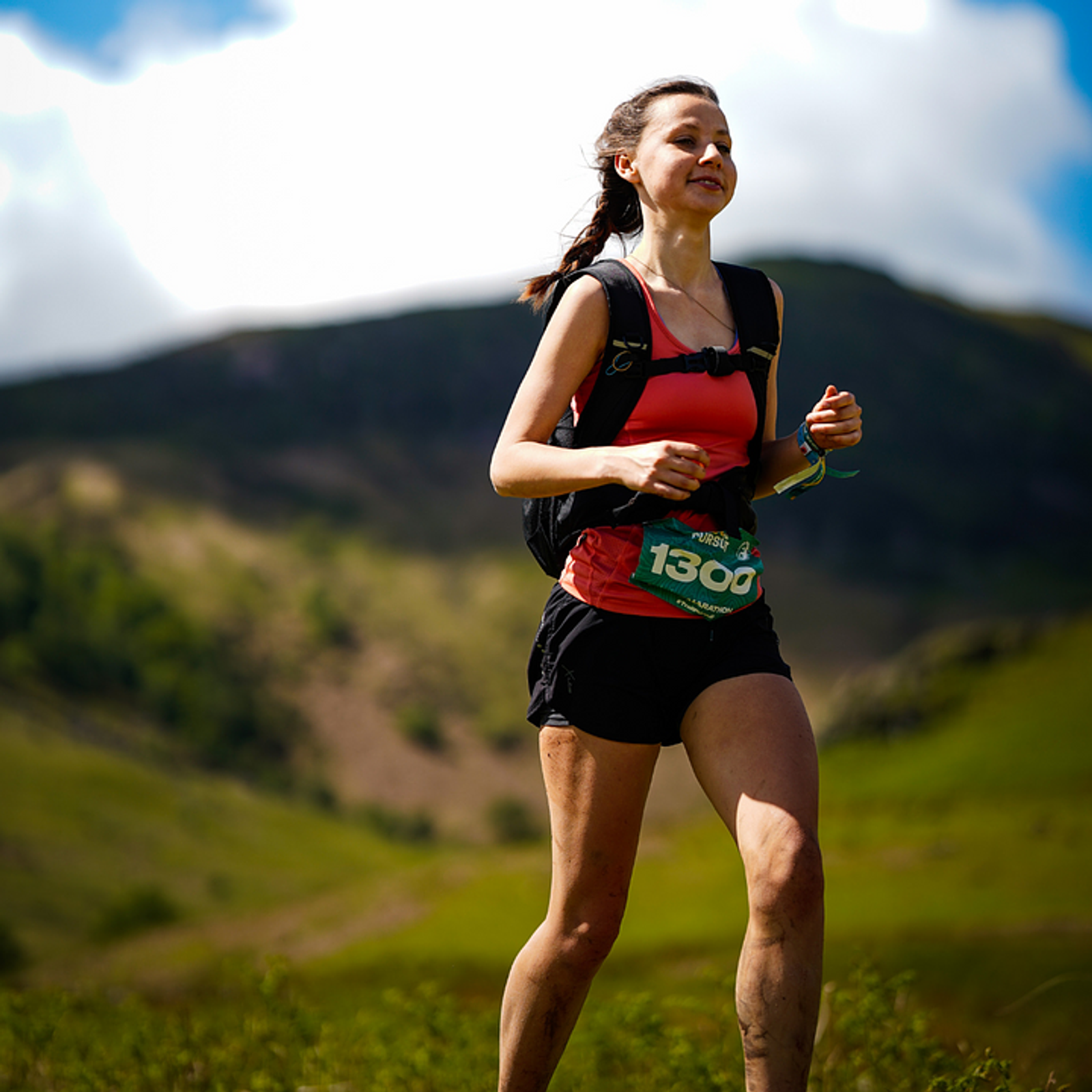 A woman in a red tank top and black shorts jogs along a trail in a green mountainous area. She wears a black backpack and a race bib with the number 1300. The background shows a clear sky with some clouds. Her legs are muddy, indicating a challenging terrain.