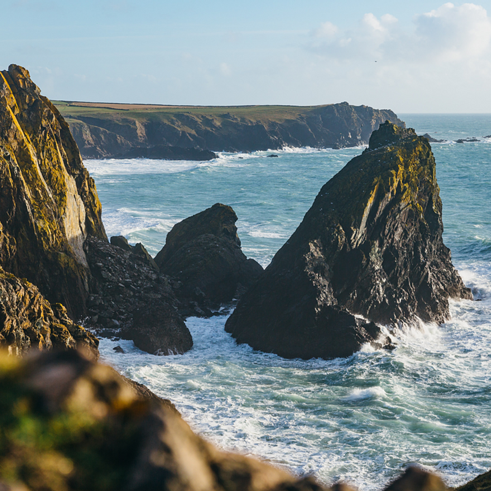 A scenic coastal view featuring rugged cliffs and large, jagged rocks jutting out from the sea. The waves crash against the rocks, and the sky is clear with a few clouds on the horizon, creating a serene and picturesque landscape.