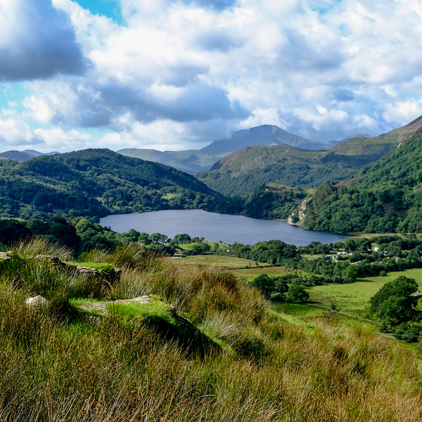 A scenic view of a lush, green valley with a serene lake in the middle, surrounded by rolling hills and mountains under a partly cloudy sky. The foreground features grassy vegetation and patches of rocks, enhancing the natural beauty of the landscape.
