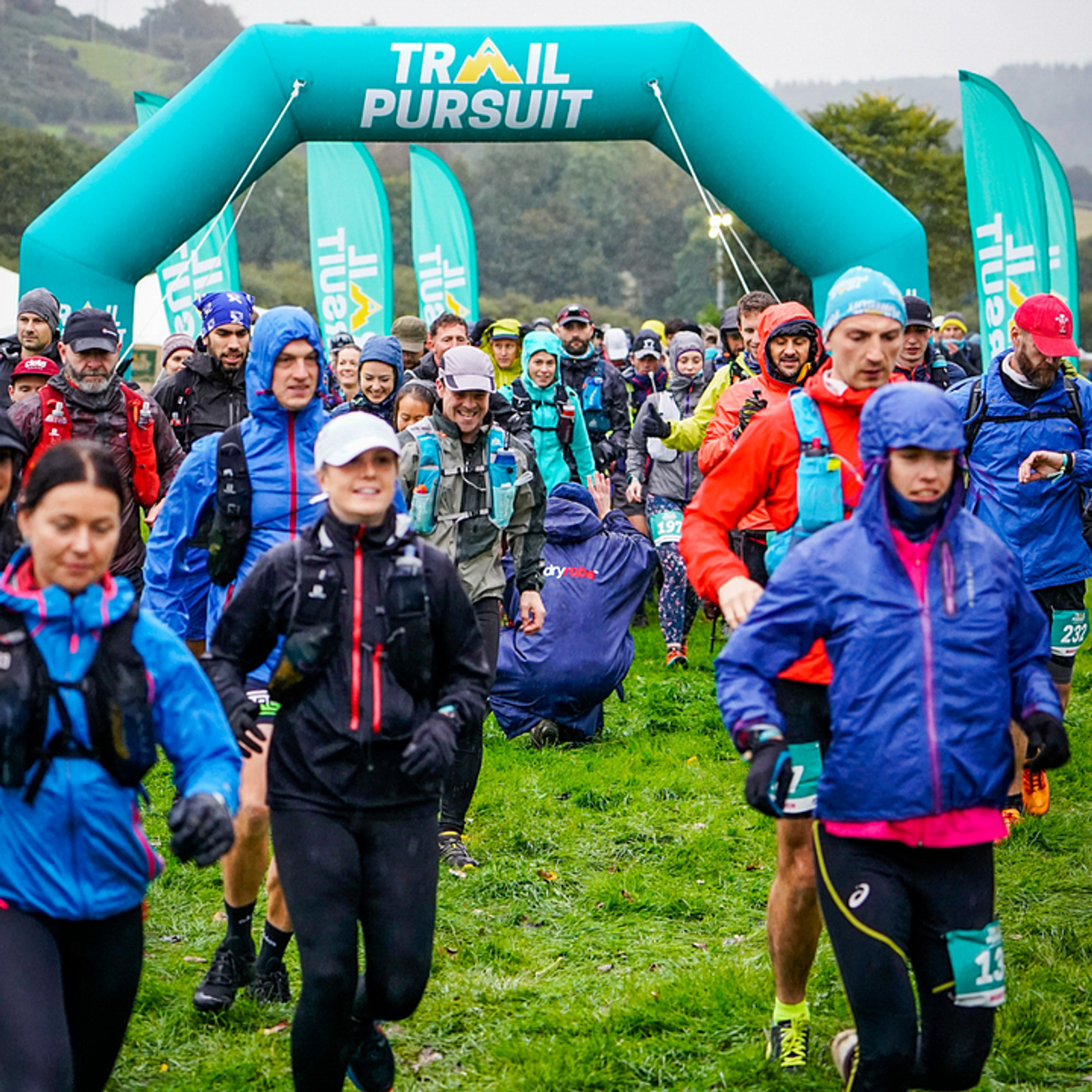 A group of runners in colorful sports attire start a trail race under an inflatable archway that reads "Trail Pursuit." They are on a grassy area with event flags in the background. The runners are of various ages, focused and determined despite the overcast weather.