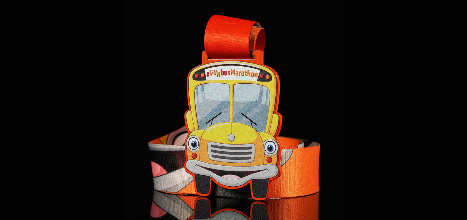 A colorful marathon medal featuring a cartoon yellow school bus with a big smile. The bus has large, expressive eyes and is labeled "FlybusMarathon" at the top. The medal is attached to a vibrant, multi-colored ribbon.