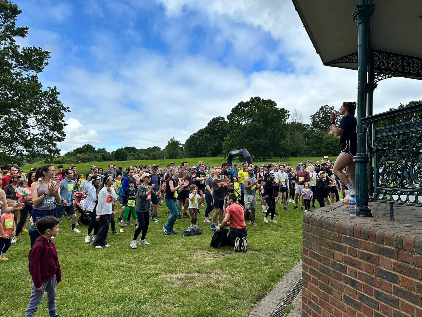 A crowd of people in athletic wear gather on grass in a park, facing a woman giving instructions from a bandstand. Some are warming up, and trees and a blue sky with clouds are in the background.