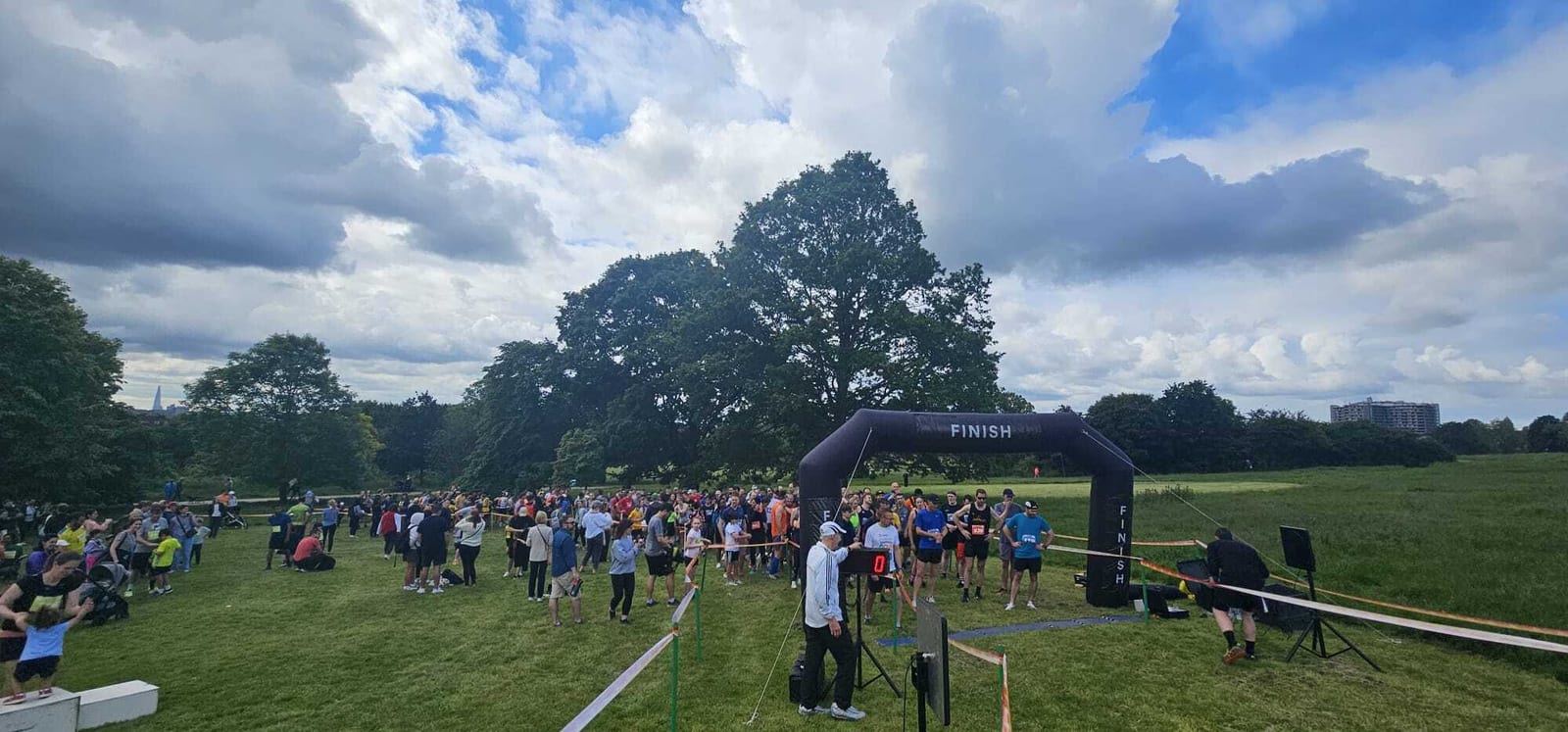 A large group of people gather at a park under a cloudy sky, some standing behind a black inflatable finish line arch, preparing for or ending a race. Trees and grassy fields surround the event.