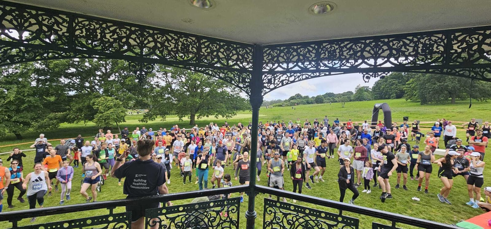 A large group of people gathers on grass for a fitness event or race, seen from a pavilion with ornate metalwork. A leader faces the crowd, and trees and green hills are visible in the background.