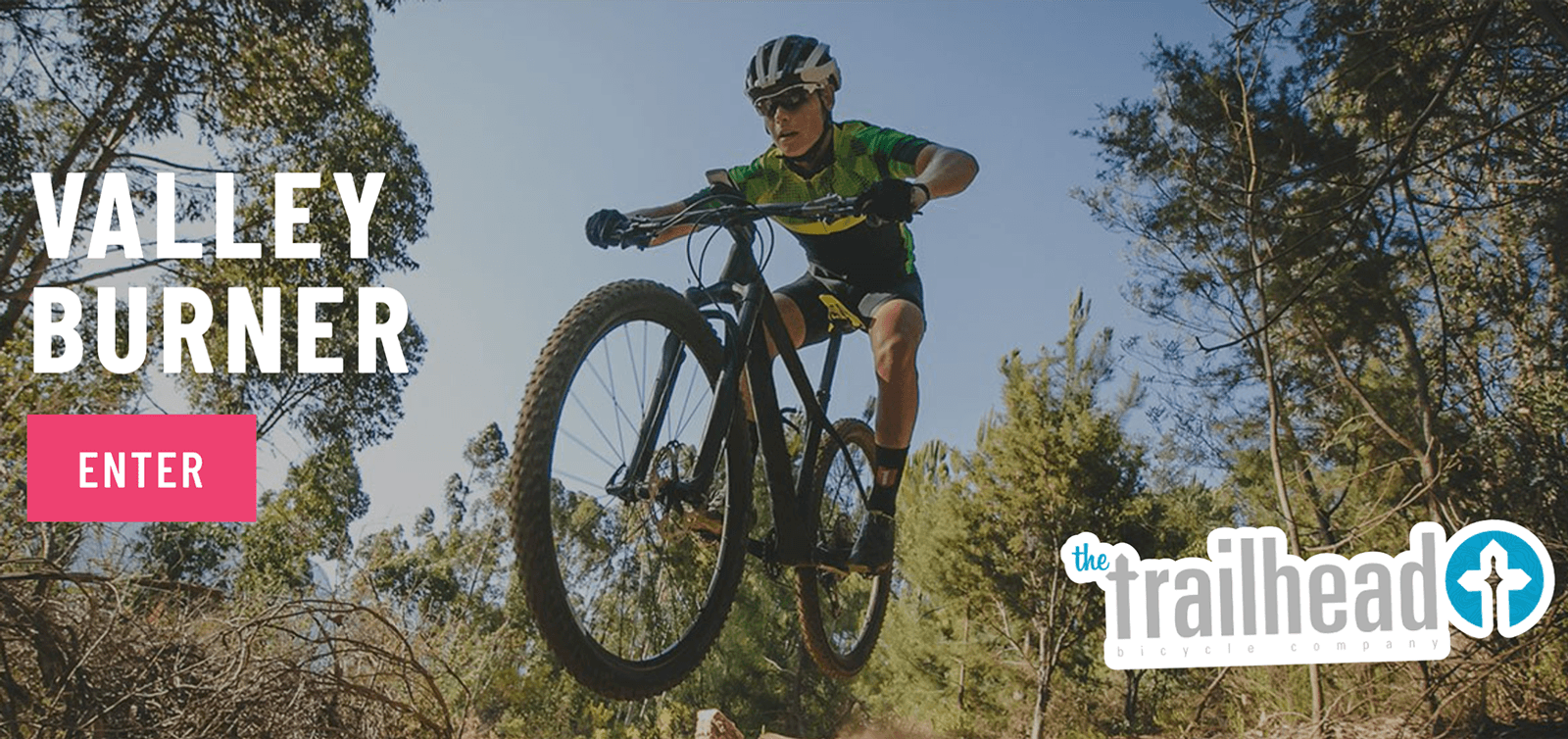 A cyclist dressed in green and black biking gear is airborne while riding a mountain bike on a trail surrounded by pine trees. Text on the left reads "Valley Burner" and "Enter" in a pink rectangular button. The Trailhead logo is in the bottom right corner.
