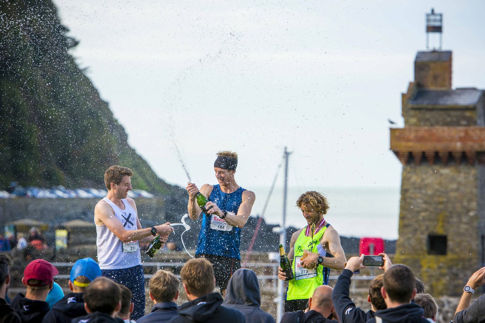 Three male runners stand on a podium, celebrating with champagne. The runner in the center is spraying champagne while the other two smile and laugh. The backdrop includes a large rock formation and stone building by the sea. It’s a sunny day with a clear sky.