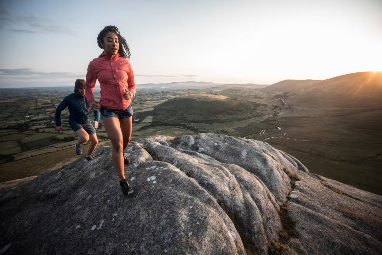 Two individuals are trail running on a rocky outcrop with a vast landscape in the background. They wear athletic gear, and the setting sun casts a warm glow over the rolling hills and plains, creating a serene and picturesque atmosphere.
