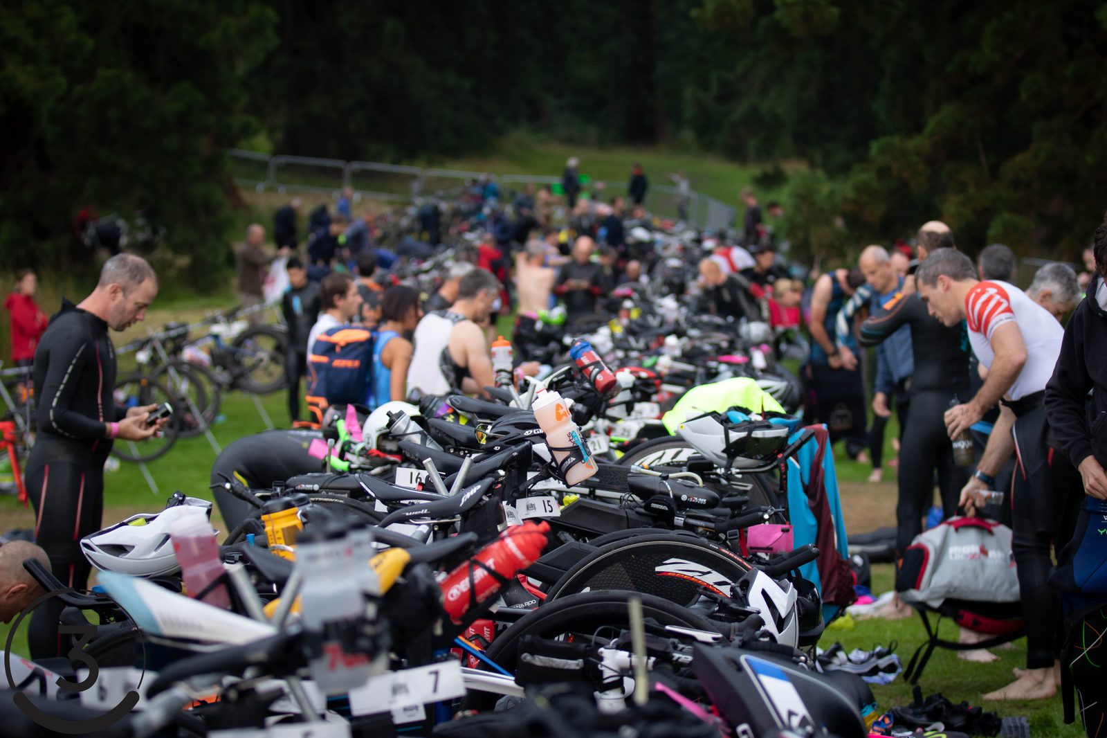 A crowded transition area at a triathlon with athletes preparing their gear. Several bicycles are racked in neat rows, and competitors are seen adjusting their equipment, putting on wetsuits, or getting ready for the next leg of the race. Trees and greenery present in the background.