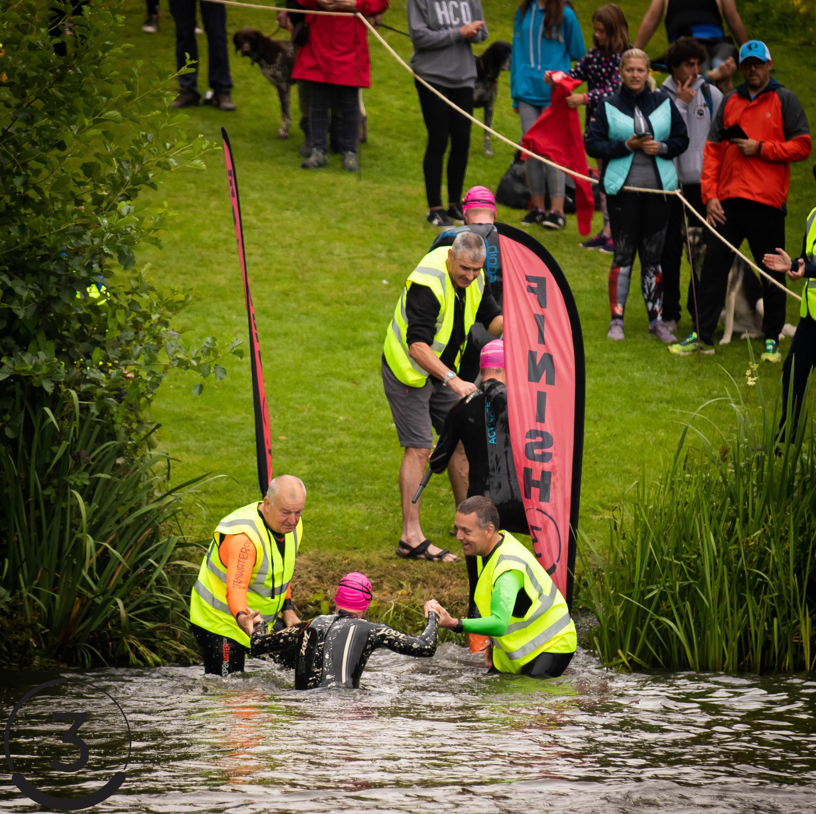 Swimmers in wetsuits and pink caps exit a lake, assisted by volunteers in yellow vests. Two large red and black flags marked "FINISH" are visible. Spectators in colorful clothing stand behind a fence, watching from the grassy shore.