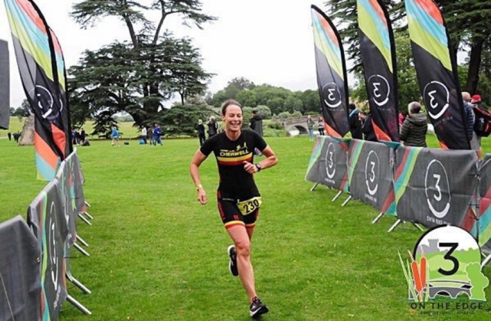 A female athlete, wearing a black and red triathlon suit with the number 129, runs towards the finish line at an outdoor race event. Colorful banners and spectators line the path. Behind her is a grassy area with large trees and a distant stone bridge.