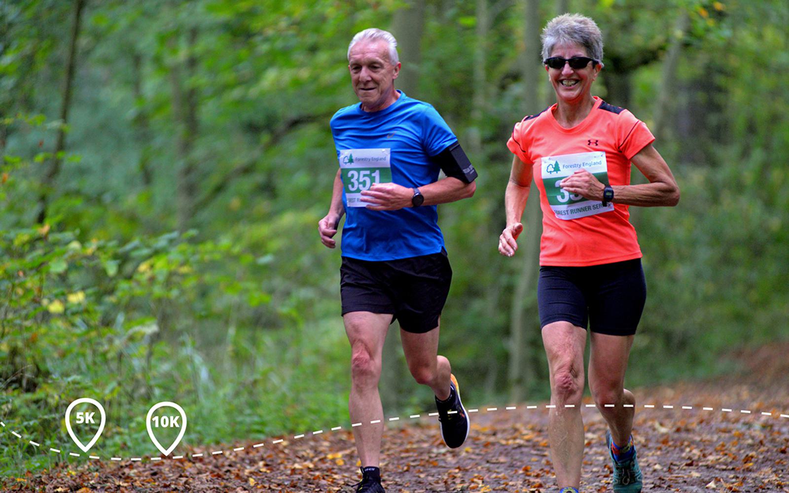 Two runners are participating in a race on a wooded trail. The older man on the left wears a blue shirt with the number 351, while the woman on the right, wearing a red shirt, sports the number 353. Both are smiling as they run amidst green foliage.