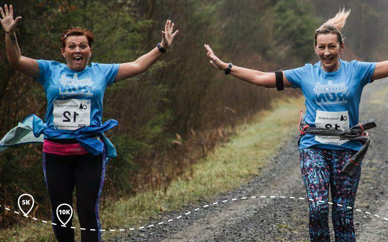 Two women running on a gravel path lined with grass and trees, both smiling and raising their arms in joy. They wear blue shirts and race bibs, with one indicating "5" and the other "8". A dotted line marks distances of 5K and 10K on the ground.